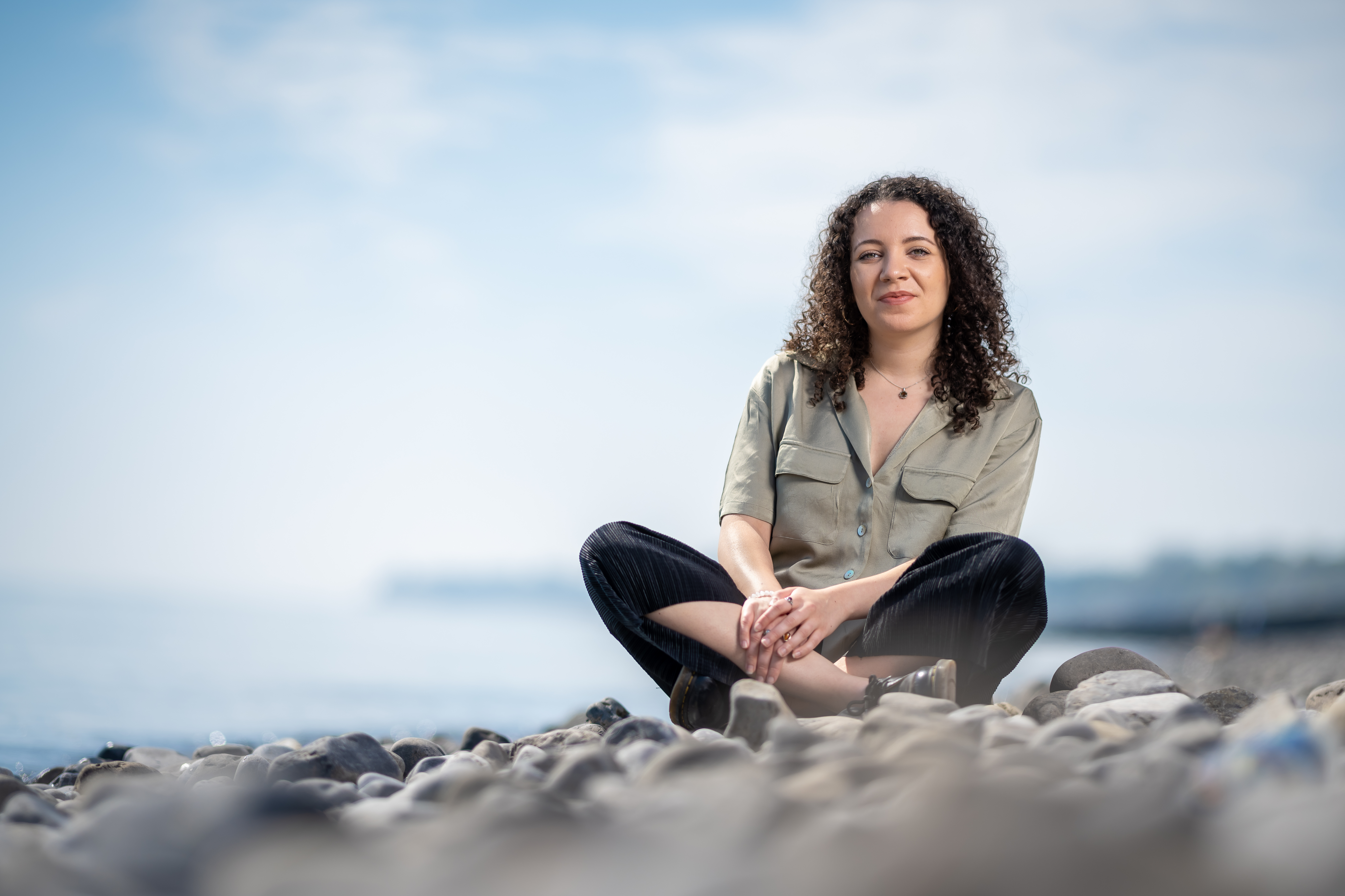 Image of the new poet in residence, sitting on a pebble beach and smiling. 