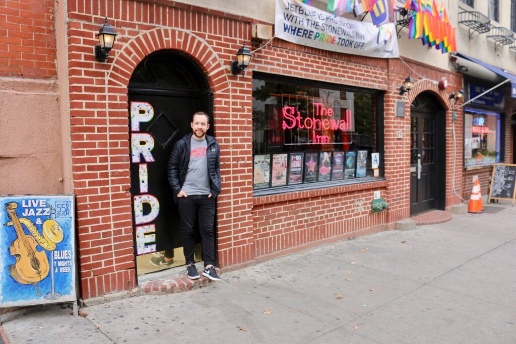 Jacob Ellis our Lead Change Maker smiling and standing in front of the Stonewall Inn which displays a sign saying 'where pride took off'. 