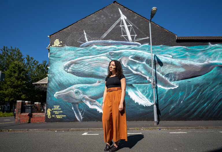 Taylor Edmonds, Bardd Preswyl Comisiynydd Cenedlaethau'r Dyfodol Cymru, wrth ymyl y murlun morfilod enfawr, a baentiwyd gan Spike Clark, yng Nghaerdydd. Mae'r gwaith celf yn tynnu sylw at gynhesu byd-eang a lefelau'r môr yn codi.