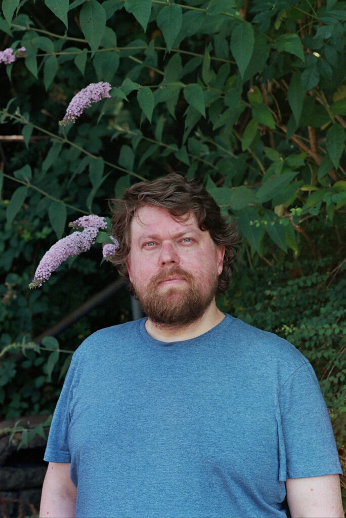 Man wearing a blue t-shirt looking directly at the camera in front of a tree. 