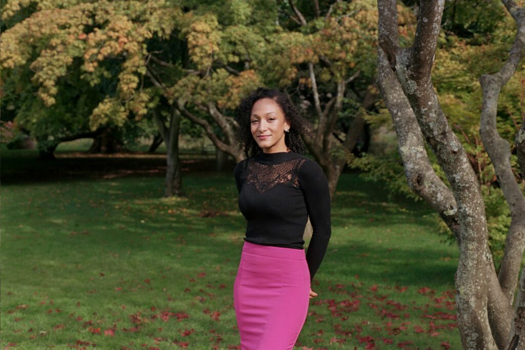 Young Black woman, Jessica Dunrod, stands in front of big green trees with her arms behind her. 