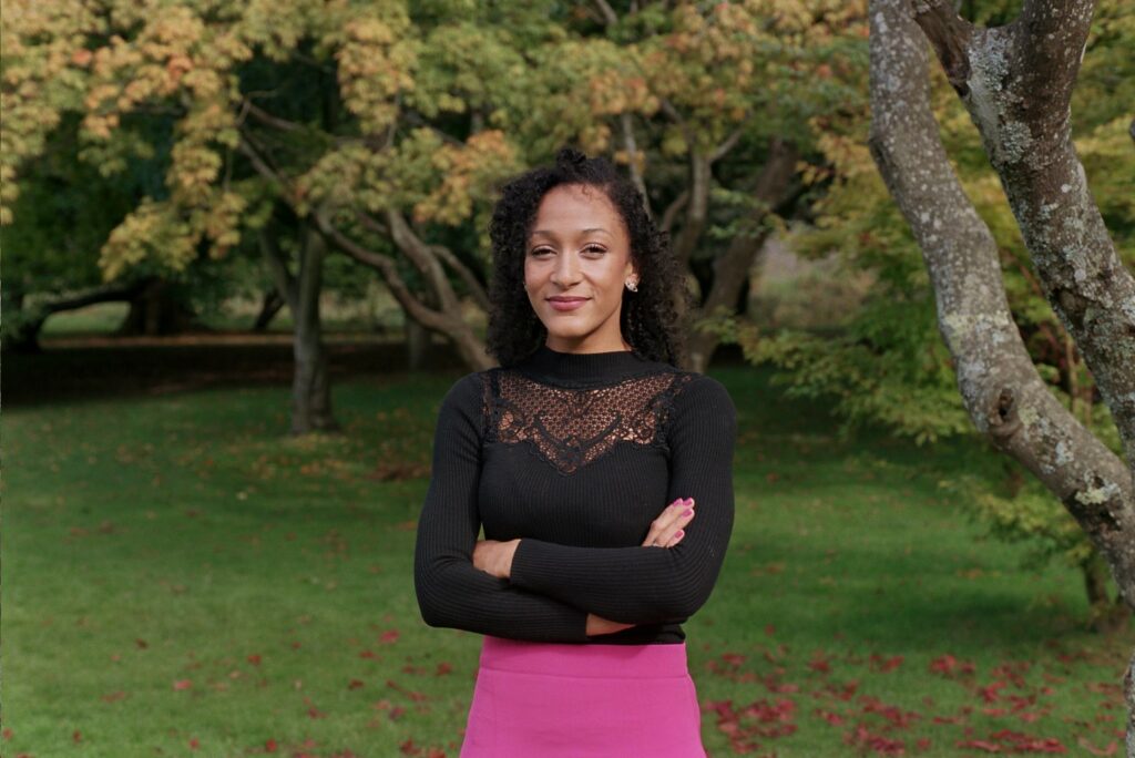 Young Black woman, Jessica Dunrod, stands in front of big green trees with her arms folded in front of her. 